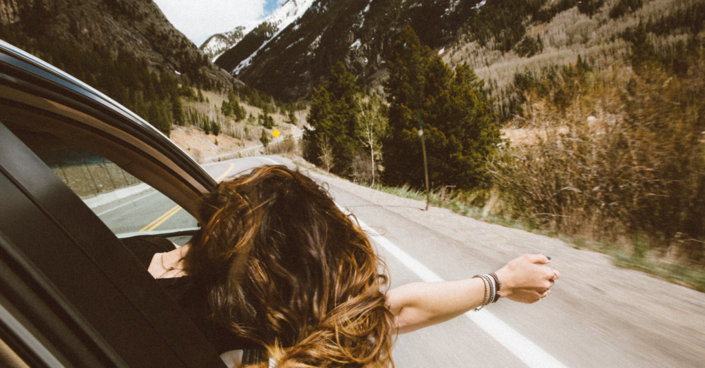 A brown haired girl driving on a scenic route with her head and hand sticking out of the car.