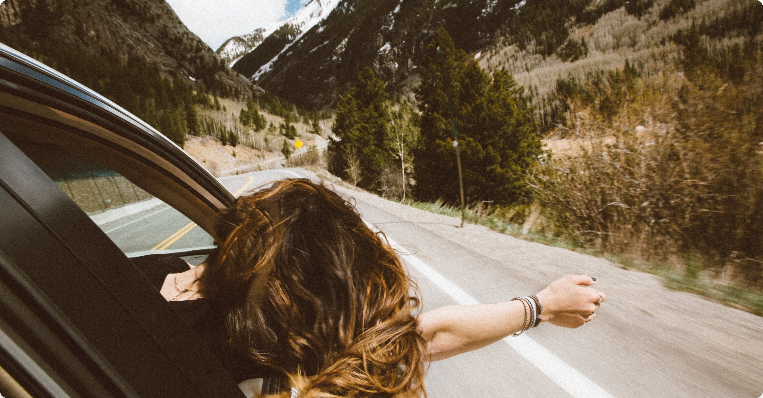 Brown haired girl driving a car with her head and right hand outside the window.