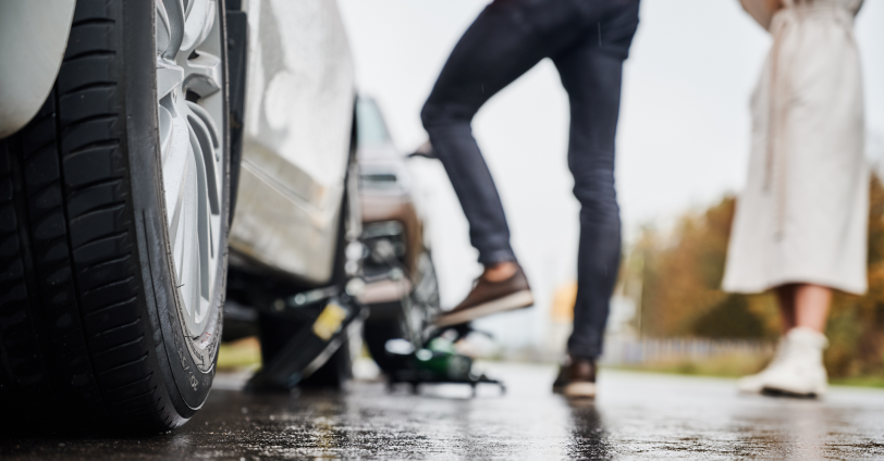 2 pairs of legs on a wet pavement, with 1 person trying to change one of the car tyres.